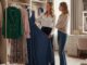 Una Personal Shopper sorridente che mostra a una cliente alcuni abiti appesi in un elegante camerino o guardaroba, con l'intento di scegliere l'abbigliamento più adatto al suo stile.
